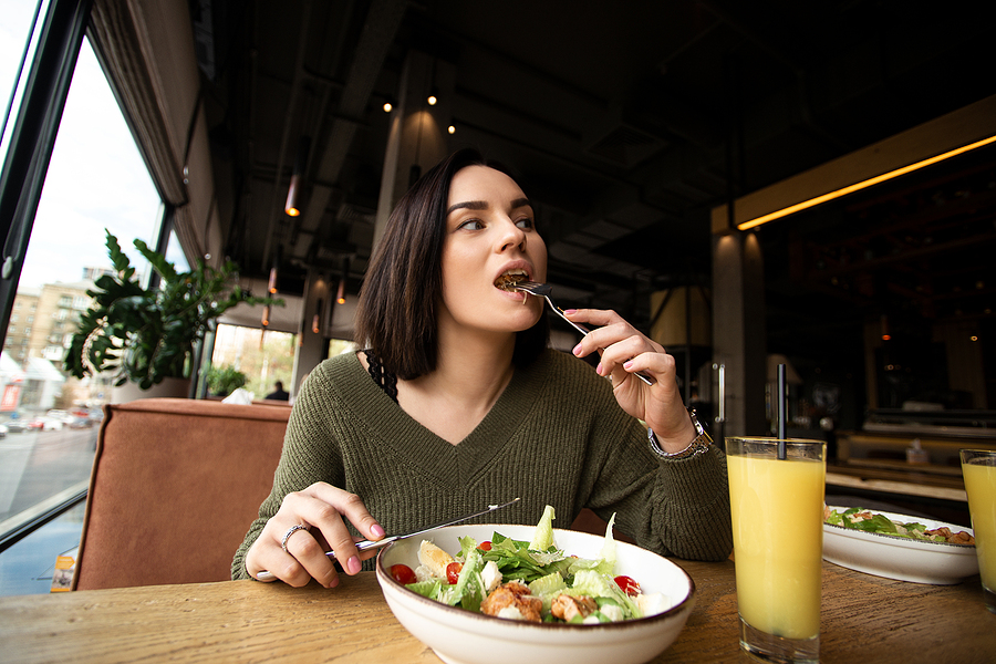 Young woman enjoys tasty meal. Attractive woman with brown hair slowly eating healthy caesar salad and looking away. Glass of orange juice. Restaurant or cafe on background. Cozy atmosphere. Miami County menu